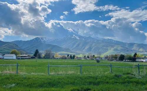 A scenic view of green fields with a wooden fence, mountains in the background, and a partly cloudy sky.