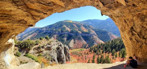 A person sits in a cave opening, overlooking a vibrant autumn landscape of mountains and trees.