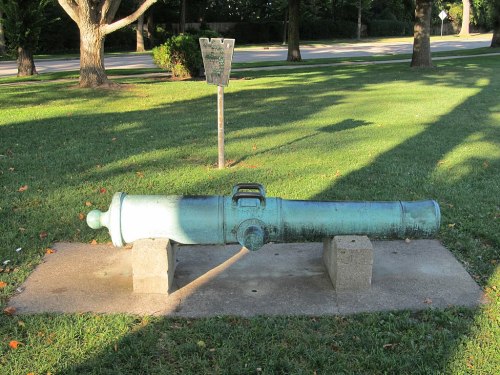 A green cannon displayed on a stone base in a grassy area, with a sign visible in the background.