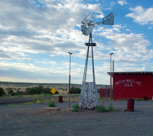 A windmill stands beside a red building with "Witch Well, AZ, U.S.A." painted on it, against a cloudy sky.