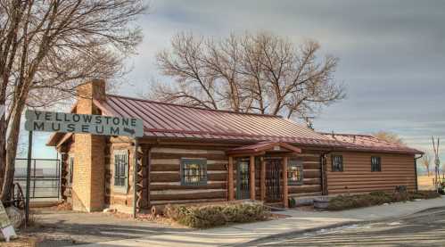 Log cabin-style building with a red metal roof, featuring a sign that reads "Yellowstone Museum" and surrounded by trees.