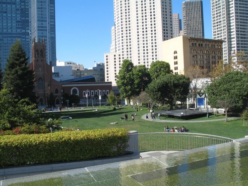 A city park with green lawns, trees, and people relaxing, surrounded by tall buildings and a church in the background.