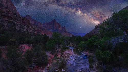 A starry night sky over a canyon with a river and trees, showcasing the Milky Way and rugged mountain peaks.