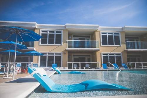 A modern pool area with blue lounge chairs and umbrellas, surrounded by a stylish building under a clear blue sky.