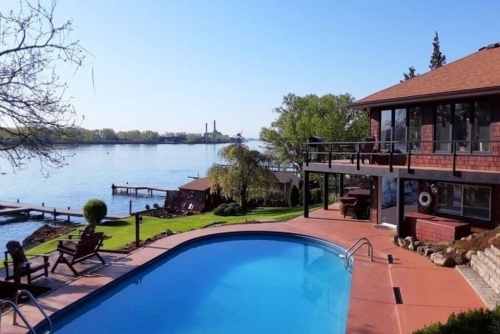 A serene lakeside view featuring a pool, lush greenery, and a house with large windows overlooking the water.