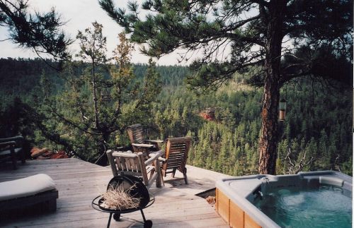 A serene outdoor deck with wooden chairs, a hot tub, and a view of lush green trees and hills in the background.