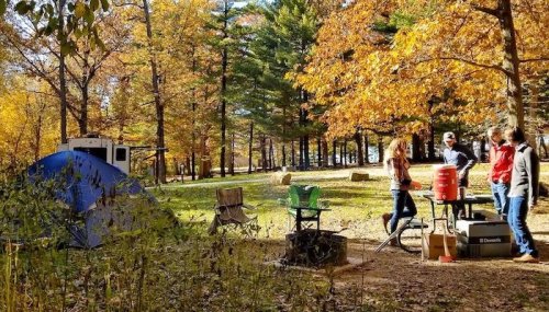 A campsite in autumn with colorful trees, a blue tent, and people gathered around a table preparing food.