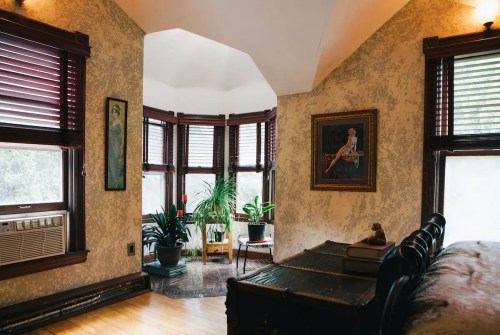 Cozy room with large windows, plants, a painting, and wooden furniture, featuring warm lighting and vintage decor.