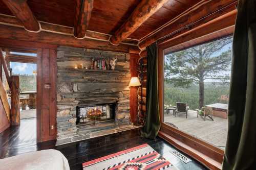 Cozy cabin interior with a stone fireplace, large window view of nature, and rustic wooden beams.