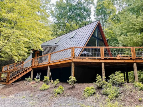 A modern A-frame cabin surrounded by trees, featuring a wooden deck and sloped roof.