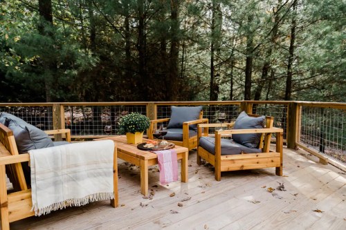 Cozy wooden deck with seating, a table, and potted plants, surrounded by lush green trees.