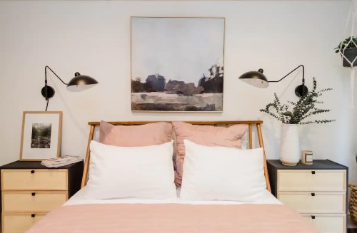 A cozy bedroom featuring a wooden bed, soft pink bedding, wall art, and stylish bedside lamps.