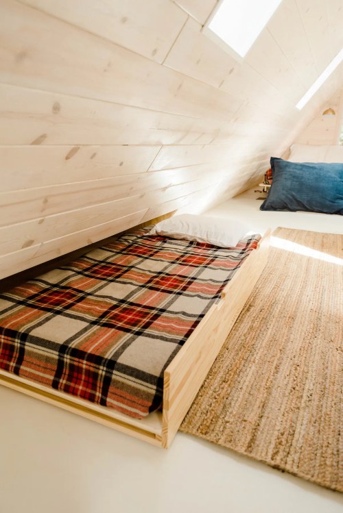 Cozy attic bedroom with a wooden bed, plaid blanket, and a soft rug, featuring sloped wooden walls and natural light.