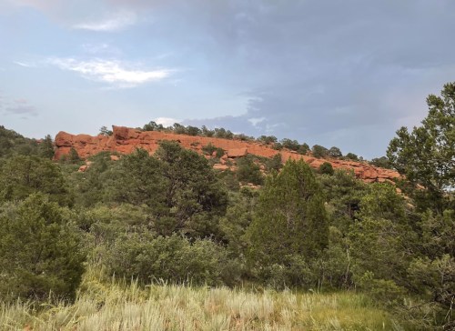 A rocky red cliff rises above a lush green landscape under a cloudy sky.