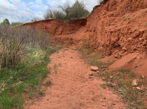 A sandy path winds through red clay cliffs, surrounded by sparse vegetation and a cloudy sky.