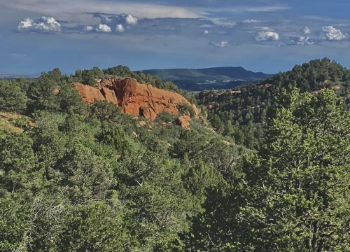 A scenic view of red rock formations surrounded by lush green trees and a blue sky with scattered clouds.