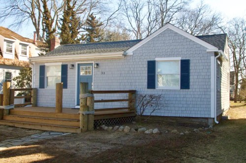 A small blue house with white trim, featuring a wooden deck and blue shutters, surrounded by trees and grass.