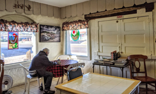 A person sits alone at a table in a cozy café, surrounded by colorful window decorations and a bookshelf.