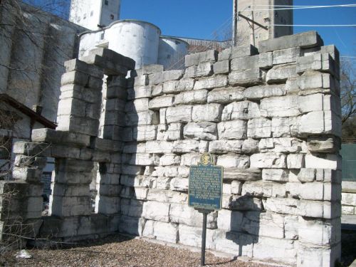 Ruins of a stone structure with a historical marker in front, set against a clear blue sky.