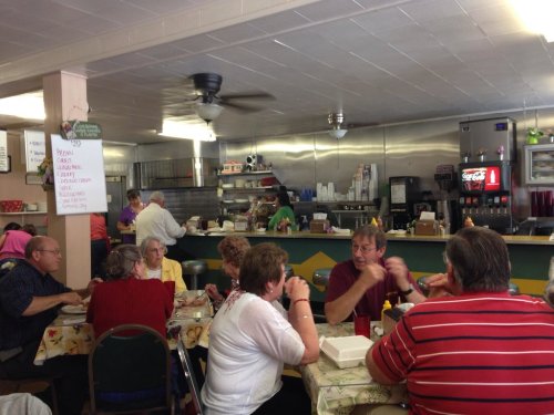 A busy diner with patrons enjoying meals at tables, a counter in the background, and a kitchen area visible.