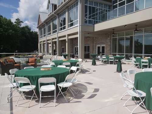 Outdoor event space with green tablecloths on round tables, white chairs, and a modern building in the background.