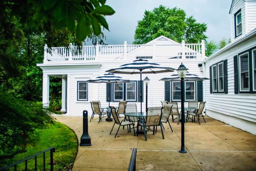 Outdoor patio with tables and chairs, shaded by umbrellas, next to a white building with a rooftop deck.