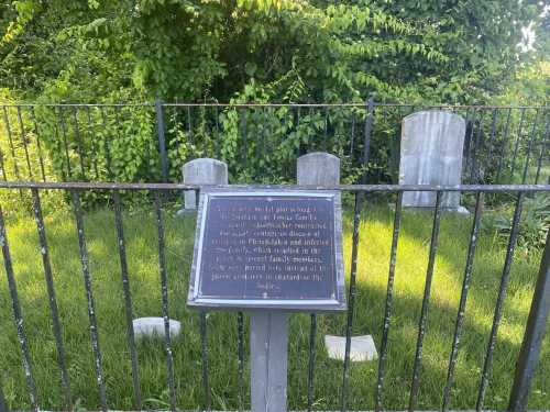 A historical marker in front of a fenced graveyard with several headstones surrounded by greenery.