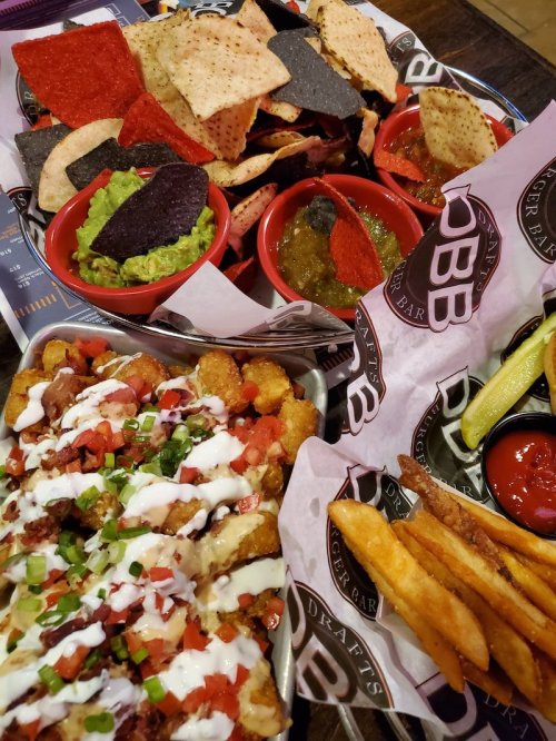 A platter of nachos, guacamole, salsa, and loaded tater tots with fries on a table at a restaurant.