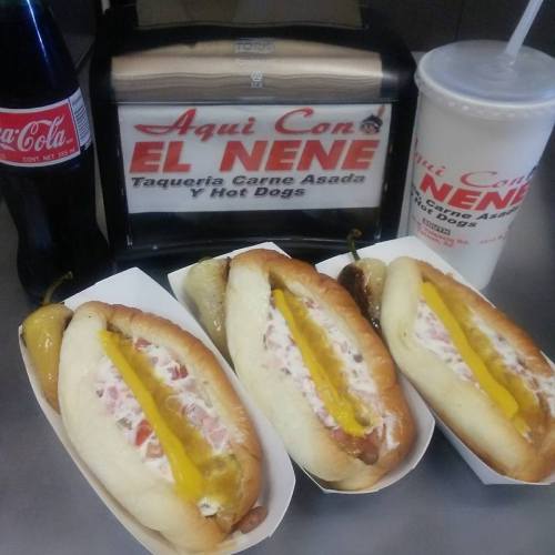 Three hot dogs with toppings in trays, a soda, and a sign for "El Nene" taqueria in the background.
