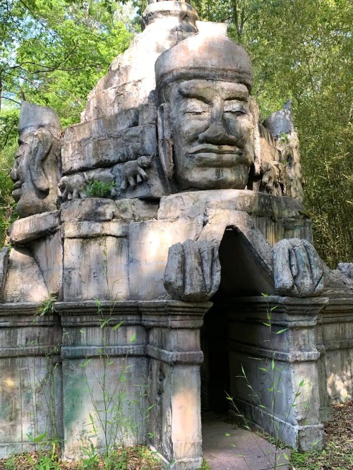 Ancient stone structure with intricate carvings and a large face, surrounded by greenery.