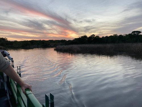 A serene sunset over a calm waterway, with colorful clouds reflecting on the surface and lush greenery in the background.