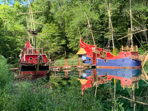 Two colorful pirate ships, one red and one blue, docked by a lush green forest, reflecting in calm water.