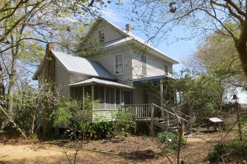 A two-story, light-colored house surrounded by trees and greenery, with a porch and a blue sky in the background.