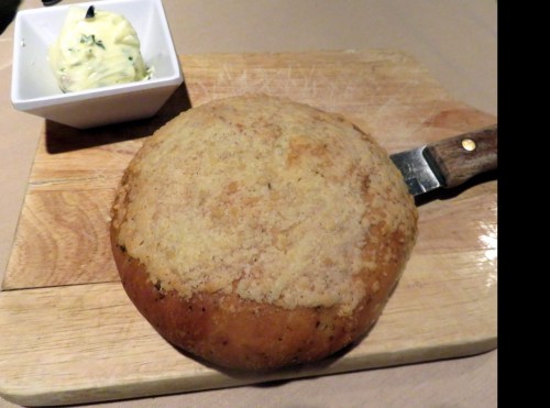 A round, crusty bread loaf on a wooden cutting board, accompanied by a small dish of butter.