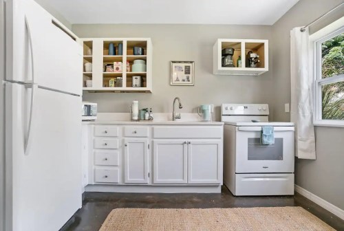 Bright kitchen with white cabinets, a stove, refrigerator, and a sink, featuring a cozy rug and natural light.