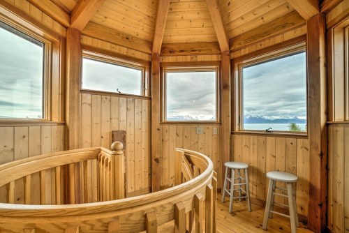 A wooden tower interior with large windows, a spiral staircase, and two stools, offering scenic mountain views.