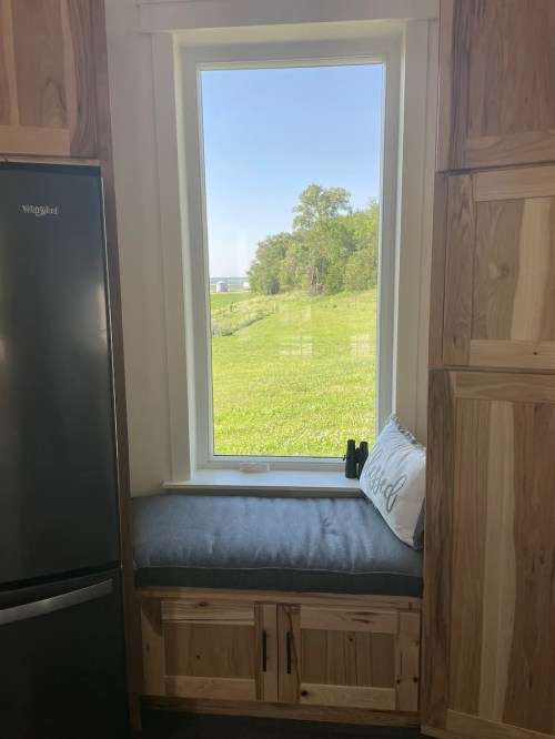 Cozy window nook with a gray cushion, surrounded by wooden cabinetry, overlooking a green landscape.