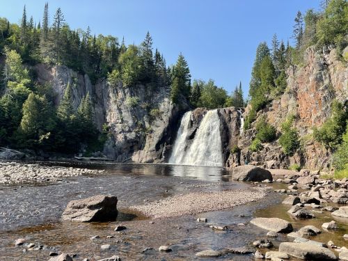A serene waterfall cascades into a calm river, surrounded by lush greenery and rocky cliffs under a clear blue sky.