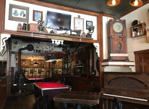 A cozy bar interior featuring a pool table, vintage decor, and a piano, with warm lighting and wooden furnishings.