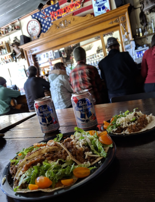 Two plates of tacos with lettuce and tomatoes, two cans of beer, and people sitting at a bar in the background.