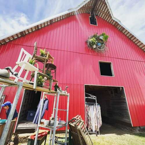 A bright red barn with a flower arrangement, surrounded by stacked chairs and clothing on a rack.