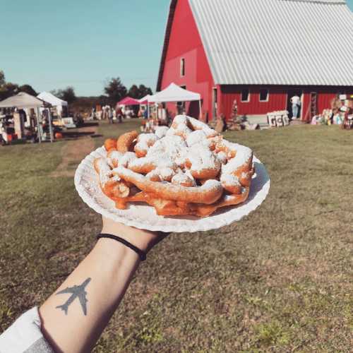 A hand holds a plate of funnel cake dusted with powdered sugar, with a red barn and market in the background.