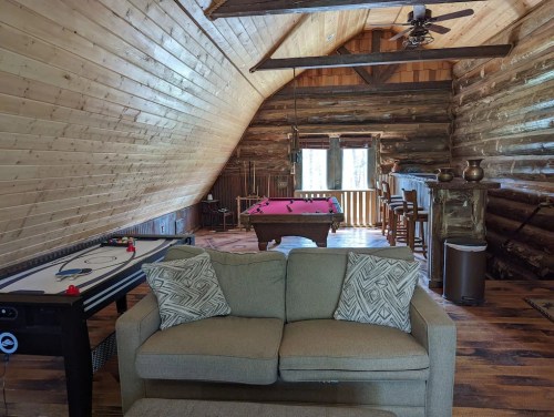 Cozy cabin interior featuring a sofa, pool table, air hockey table, and wooden walls with large windows.