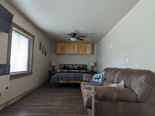 Cozy room featuring a bed, brown couch, ceiling fan, and wooden accents, with natural light from a window.