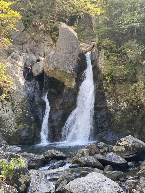 A serene waterfall cascading over rocky cliffs into a clear pool, surrounded by lush greenery and boulders.