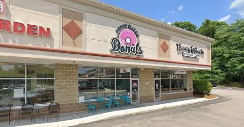 Exterior view of a donut shop with a pink sign, outdoor seating, and a coffee shop next door on a sunny day.