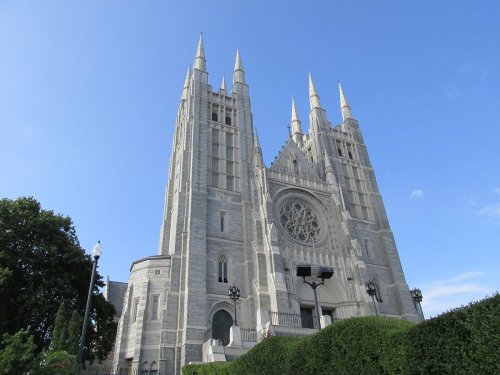 A tall, ornate stone cathedral with spires and a large rose window, surrounded by greenery under a blue sky.
