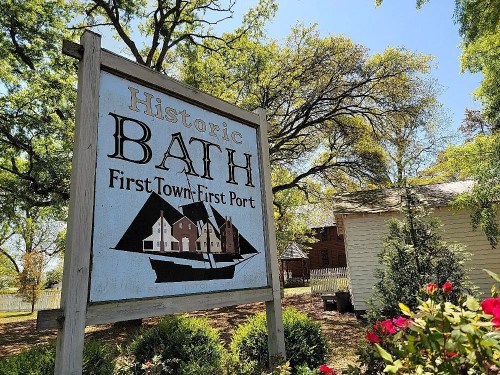 Sign for Historic Bath, featuring a boat graphic and text about the town's history, surrounded by greenery and flowers.