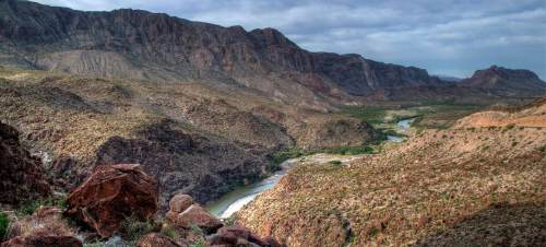 A panoramic view of a rugged landscape with mountains, a winding river, and sparse vegetation under a cloudy sky.