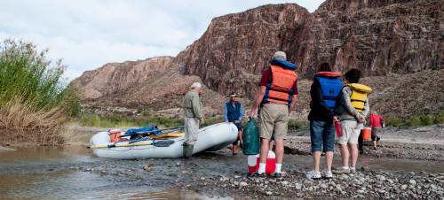 A group of people in life jackets stands by a raft near a river, surrounded by rocky cliffs and greenery.